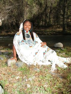 a woman sitting on the ground next to a river with rocks and grass around her