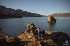 two people standing on rocks near water with mountains in the background