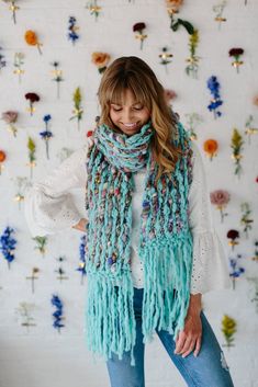 a woman standing in front of a flower wall wearing a blue scarf with fringes