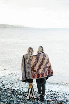 two women standing on the beach with their back to each other, wrapped in a blanket