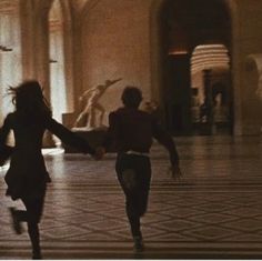 two people are running in the middle of a room with statues on either side of them