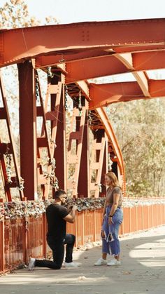 a man taking a photo of a woman sitting on a bench in front of a bridge