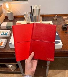 a person holding two red books in front of a desk with many other items on it