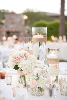 a table topped with lots of vases filled with white and pink flowers next to candles