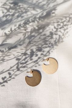 two pairs of gold earrings sitting on top of a white cloth covered table next to a plant