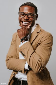 a man wearing glasses and a tan jacket smiles at the camera with his hand on his chin