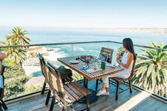 a woman sitting at a wooden table on top of a balcony next to the ocean