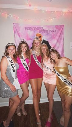 several women in dresses and tiaras posing for a photo with a man on the wall behind them