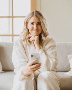 a woman sitting on a couch holding a cell phone and looking at the camera while smiling