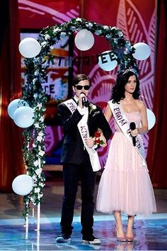 a man and woman standing on stage with microphones in front of an arch decorated with balloons