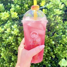 a person holding up a pink drink with a yellow straw in their hand and green bushes behind it