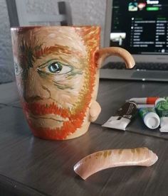 a cup with a face painted on it next to some paintbrushes and a laptop