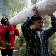 two men carrying large bags on their heads in the woods, one man is wearing a red jacket and the other wears a plaid shirt