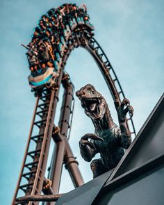 a statue of a dinosaur on top of a roller coaster at an amusement park with people in the background