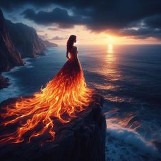 a woman standing on top of a cliff next to the ocean with fire in her dress