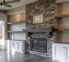 an empty living room with stone fireplace and built - in shelving units, windows, and ceiling fan