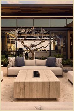 Elegant outdoor lounge area with a large concrete coffee table, plush sofas, decorative pillows, and a bare tree centerpiece, under a modern wooden pergola. Coastal Design, Honeymoon Destinations