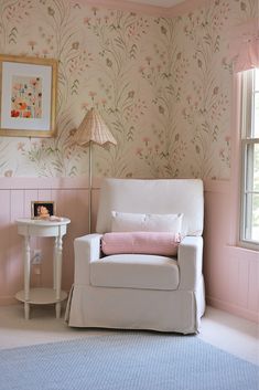 a white chair sitting in front of a window next to a table with a lamp on it