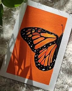 an orange and black butterfly sitting on top of a piece of paper next to a plant