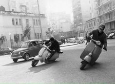 two people riding scooters in the middle of an old street with cars and buildings