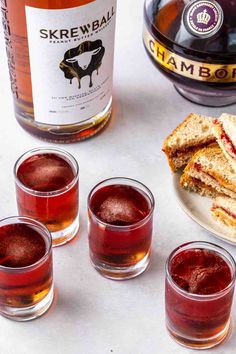 three glasses filled with drinks next to two sandwiches and a bottle of wine on a table