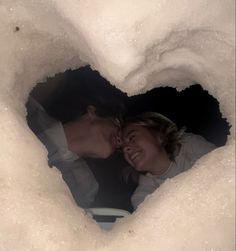 a man and woman are looking into a heart shaped hole in the snow with their faces close to each other