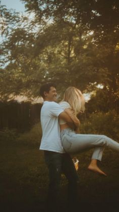 a man holding a woman in his arms while they are walking through the grass with trees behind them