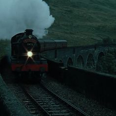 a steam train traveling down tracks next to a lush green hillside on a foggy day