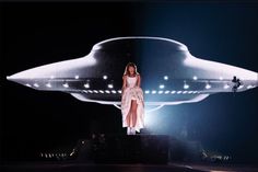 a woman standing on top of a stage in front of a giant object that looks like a flying saucer