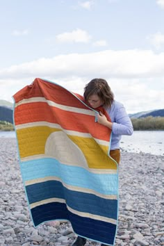 a woman is holding up a colorful blanket on the beach with rocks and water in the background