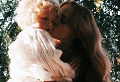 a woman holding a baby in her arms and kissing the child's face with trees in the background