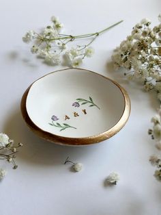 a white and gold bowl sitting on top of a table next to some wildflowers