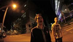 two young men standing next to each other on a street at night time, one looking up