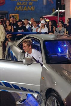 a man driving a silver sports car in front of a group of people on the street