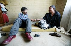 two men sitting on the floor talking to each other with shoes scattered around them and a brick wall in the background