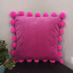 a pink pillow with pom - poms sitting on top of a table next to a potted plant