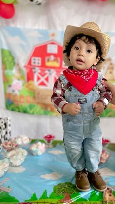 a little boy wearing overalls and a cowboy hat standing in front of a cake