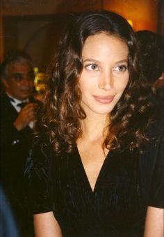 a woman with long curly hair wearing a black dress and looking at the camera while standing next to other people