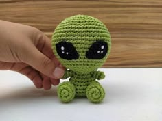 a small crocheted green alien sitting on top of a white table next to a person's hand