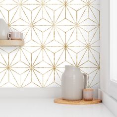 a white vase and some cups on a shelf in front of a wall with gold geometric designs