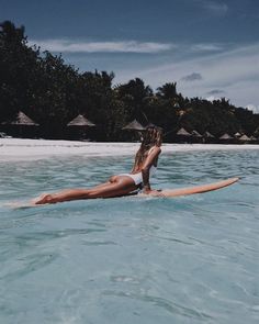 a woman is laying on a surfboard in the blue water near some huts and palm trees