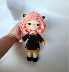 a crocheted doll with pink hair and black clothes is held up by someone's hand