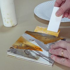 two hands are holding a piece of paper over a book on a table next to a candle