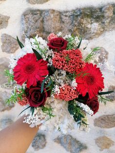 a bouquet of red and white flowers is being held by someone's hand in front of a stone wall