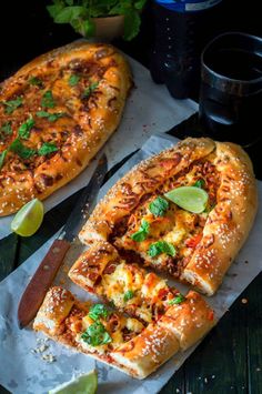 two large pizzas on top of paper next to a knife and some lime wedges