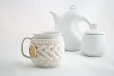 two coffee mugs and a teapot on a white surface with one being knitted