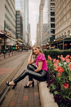 a woman is sitting on a ledge in front of some flowers and buildings while talking on her cell phone