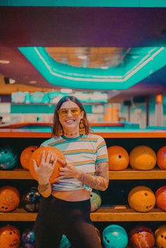 a woman holding an orange ball in front of bowling balls on the floor and shelves