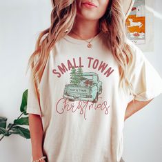 a woman wearing a small town christmas t - shirt standing in front of a potted plant