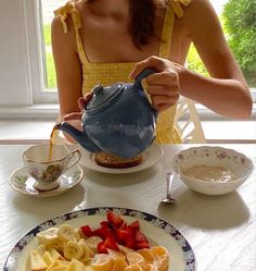 a woman in yellow dress pouring tea from a blue teapot into a plate with sliced bananas and strawberries